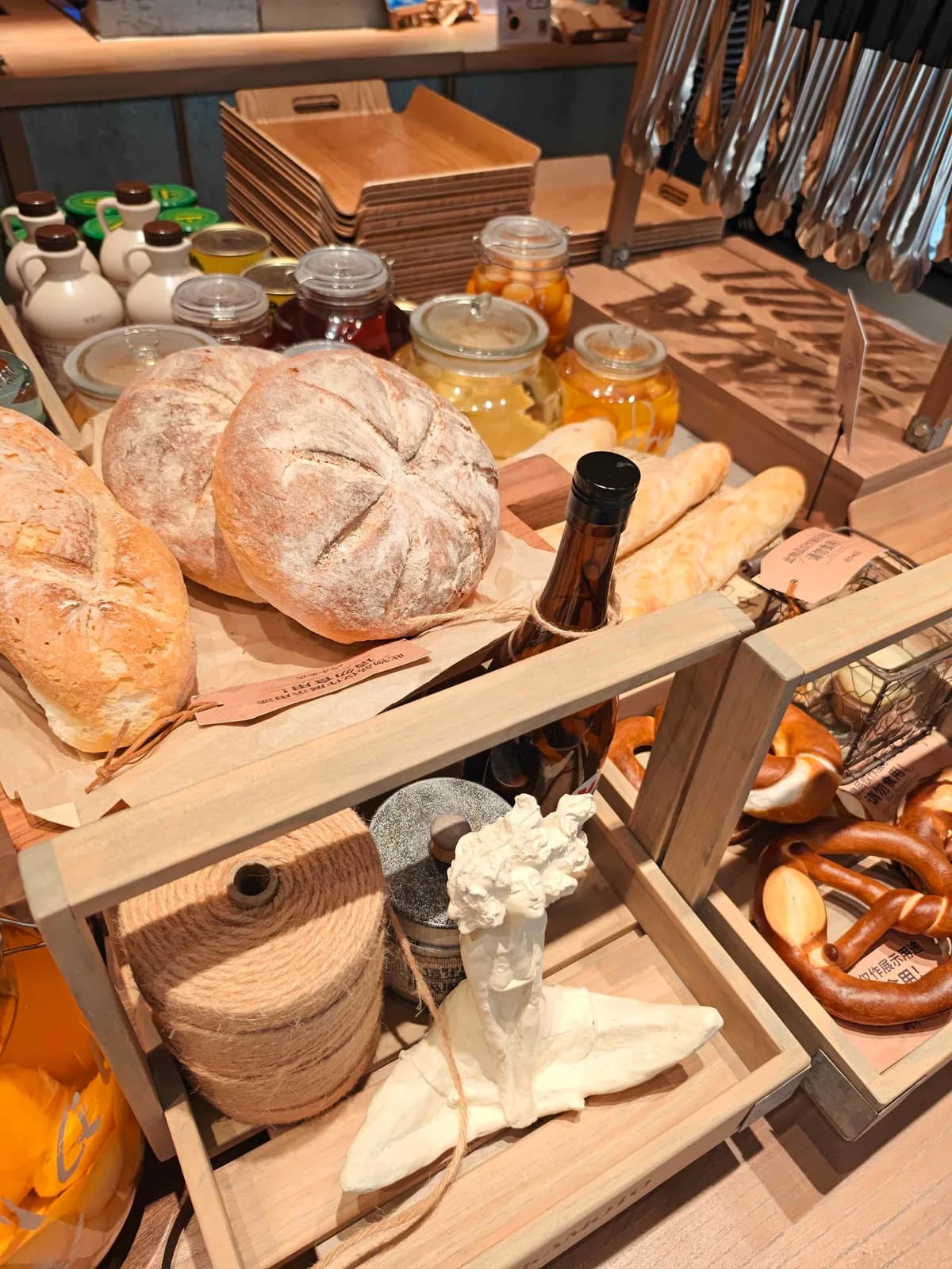 A bakery display of scored sourdough rounds and baguettes on wooden crates, with syrup jugs, honey jars and soft pretzels behind.