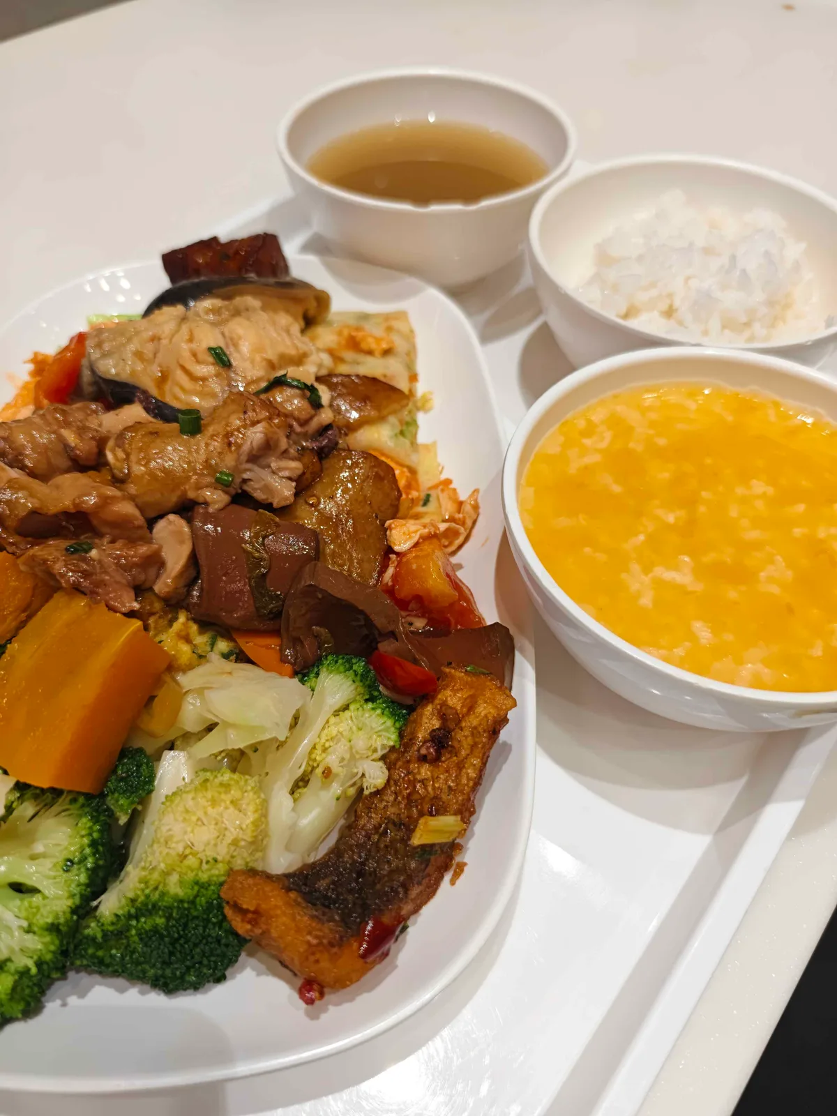 A canteen tray holding braised chicken with aubergine, broccoli and carrot over rice, with clear soup and a bowl of egg-drop congee.