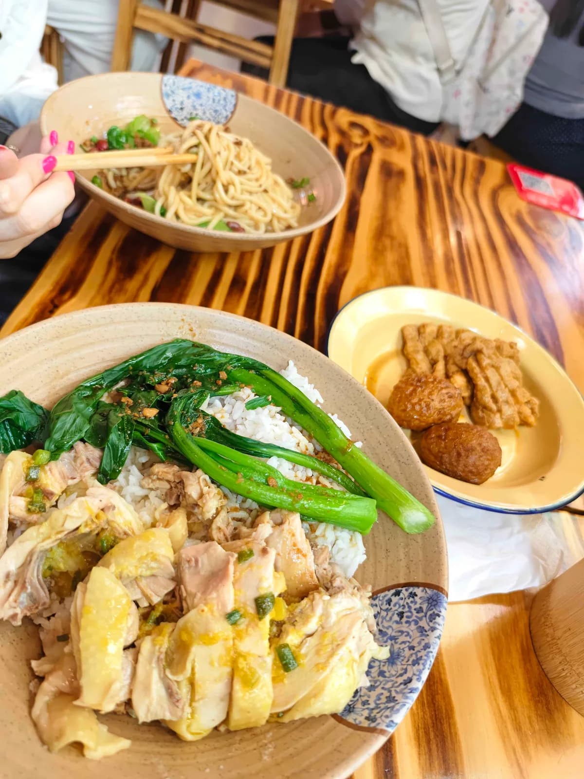 A burnt-wood table with a bowl of poached chicken and rice, a bowl of noodles, and a side plate of fried tofu skin and meatballs.