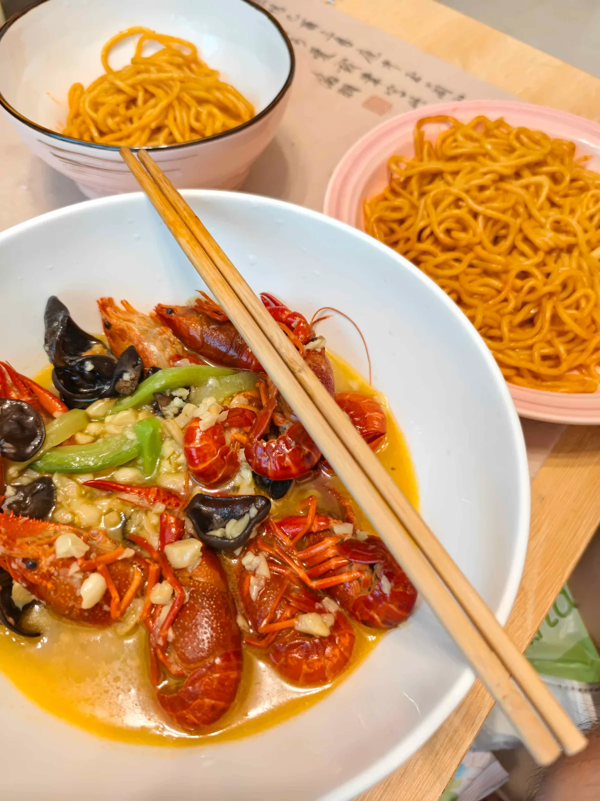 A white bowl of crayfish in garlic broth with green chilli and wood-ear mushroom, beside two plates of dark stir-fried noodles.