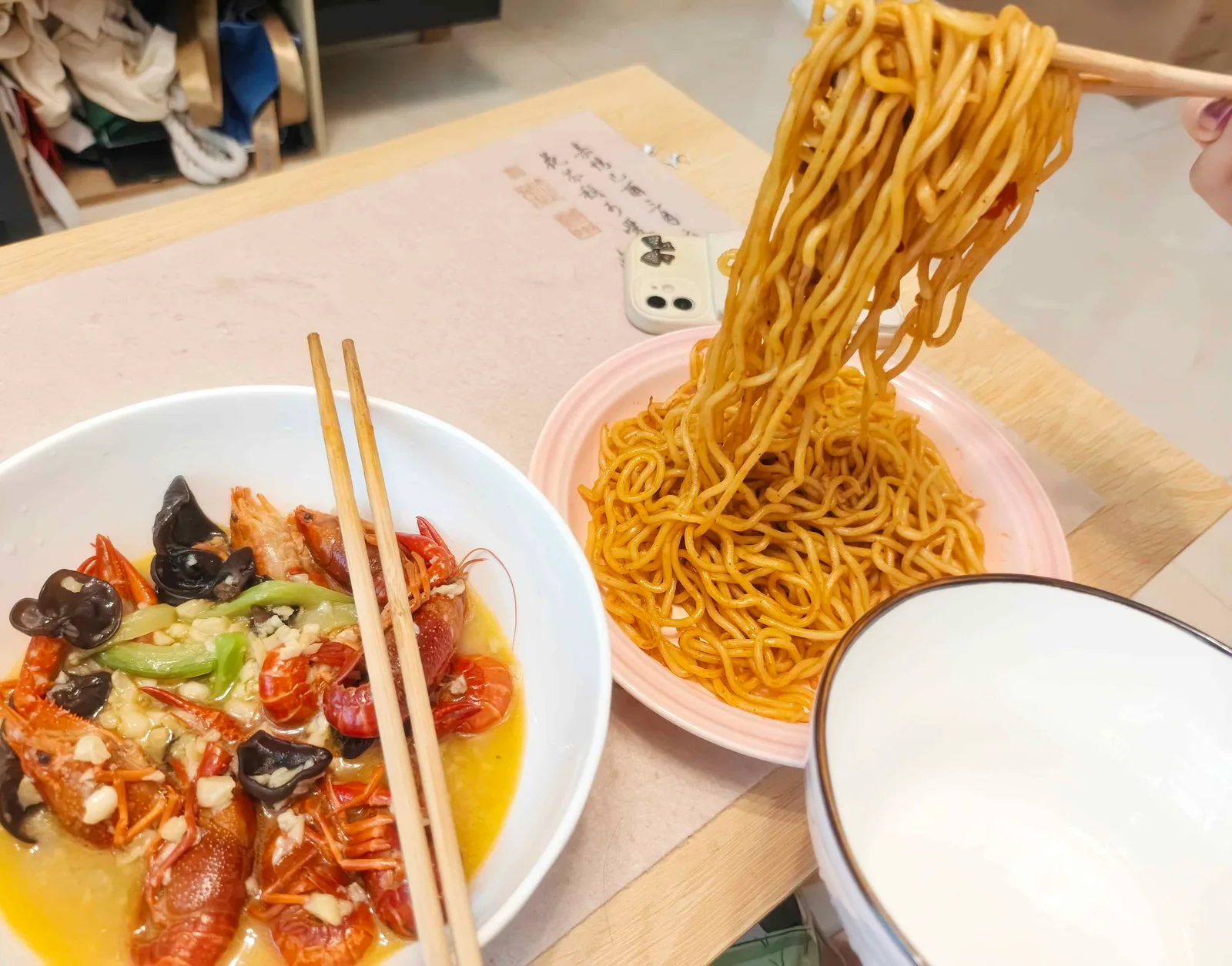 Chopsticks lifting a tangle of dark noodles from a pink plate, next to a bowl of garlic crayfish with green chilli and wood-ear mushroom.