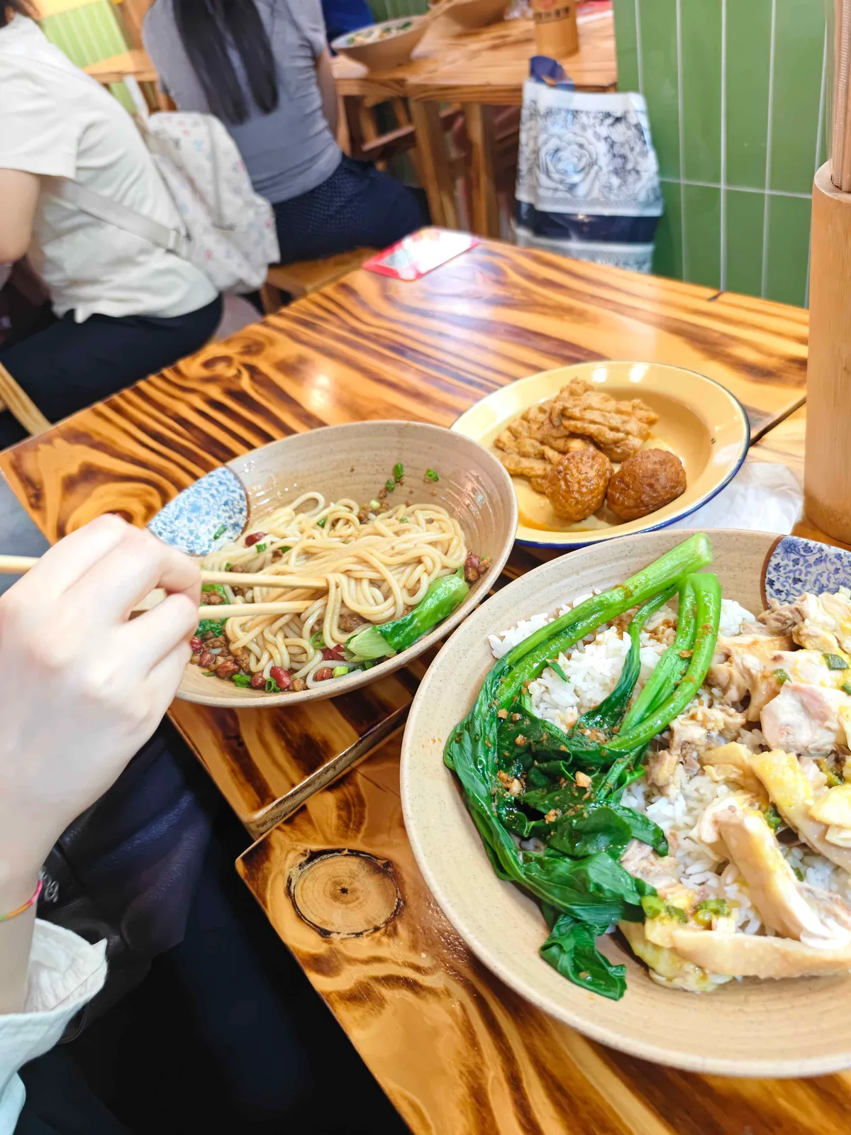 A busy noodle shop table with a bowl of noodles in chilli oil, a plate of chicken and rice with greens, and fried tofu skin.