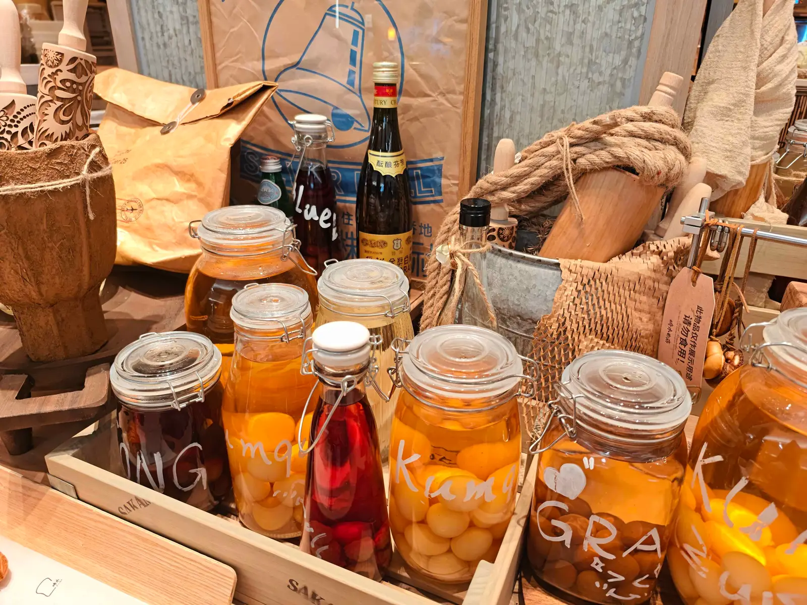 A wooden crate of clip-top glass jars holding kumquats, plums and other fruit preserved in amber and red syrup, labelled by hand.