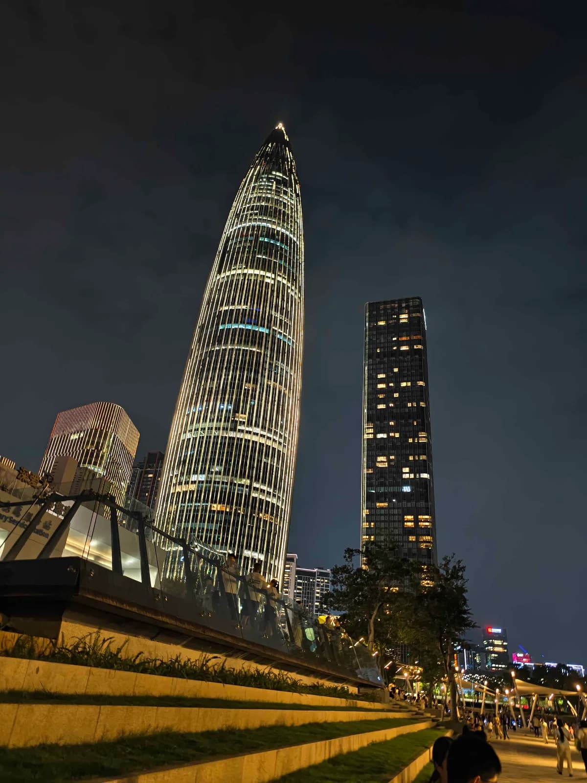 A tapered skyscraper lit white from base to spire at night, seen past a raised pedestrian walkway and a grass bank.