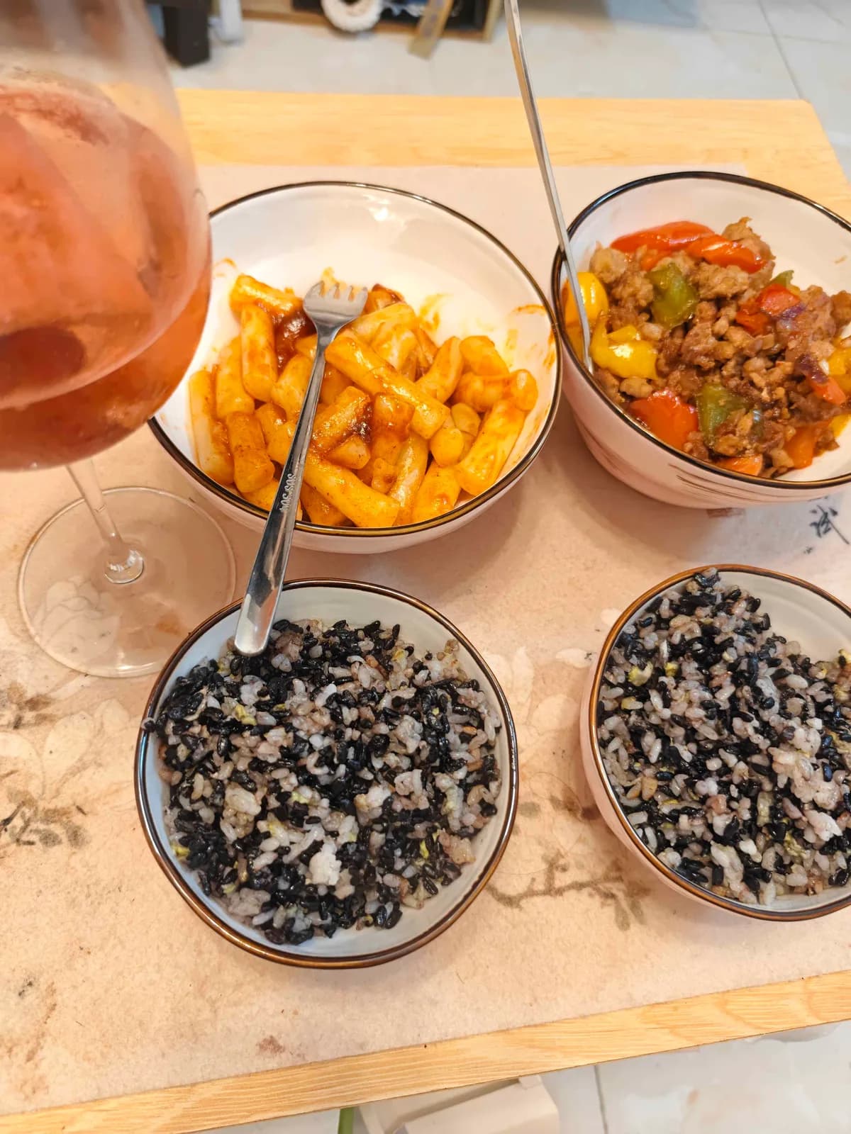 Two bowls of purple-black rice beside tteokbokki rice cakes in red sauce and a bowl of stir-fried pork with peppers.