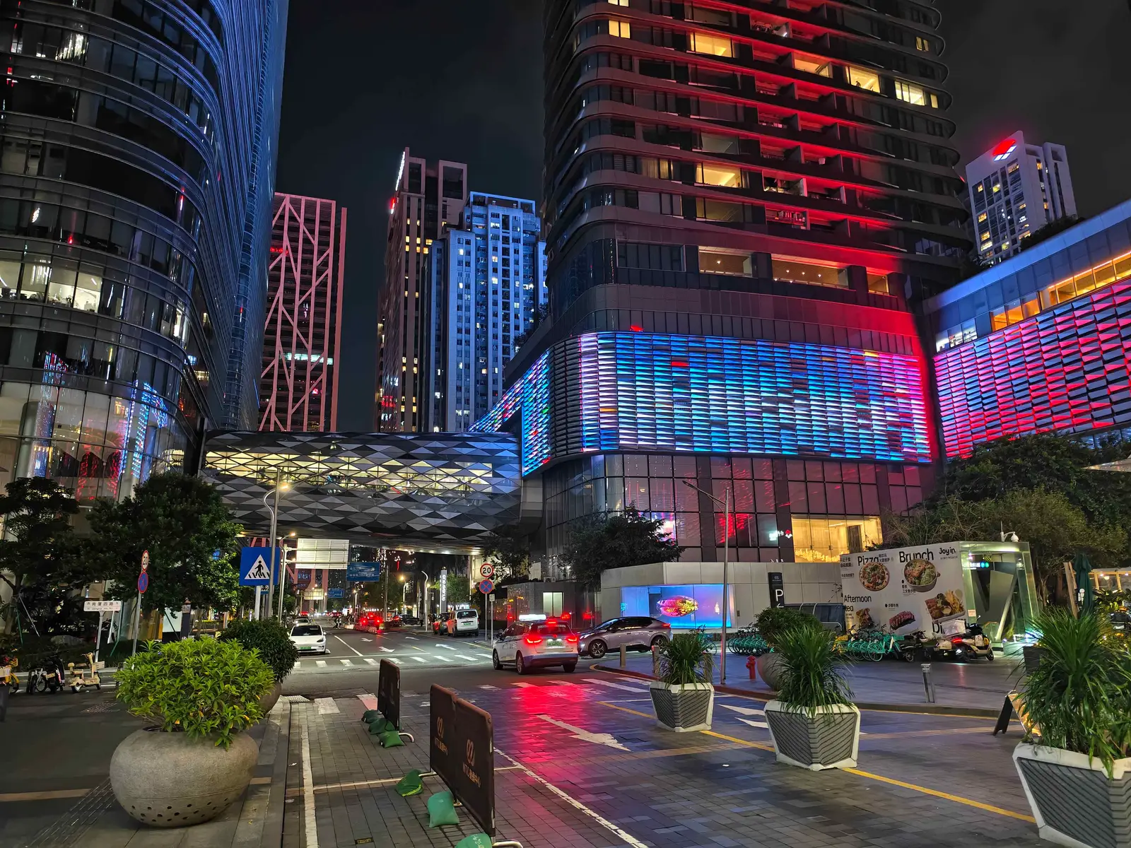 A Futian CBD street at night, towers and a mall facade lit red and blue above a glass skybridge
