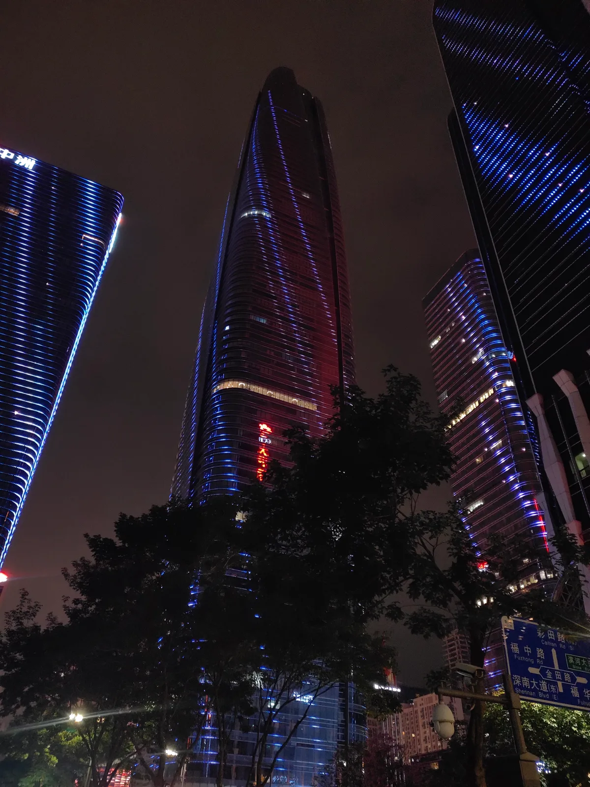 Three high-rise towers at night, two of them ribbed with blue vertical lighting, framed by dark tree branches from street level.